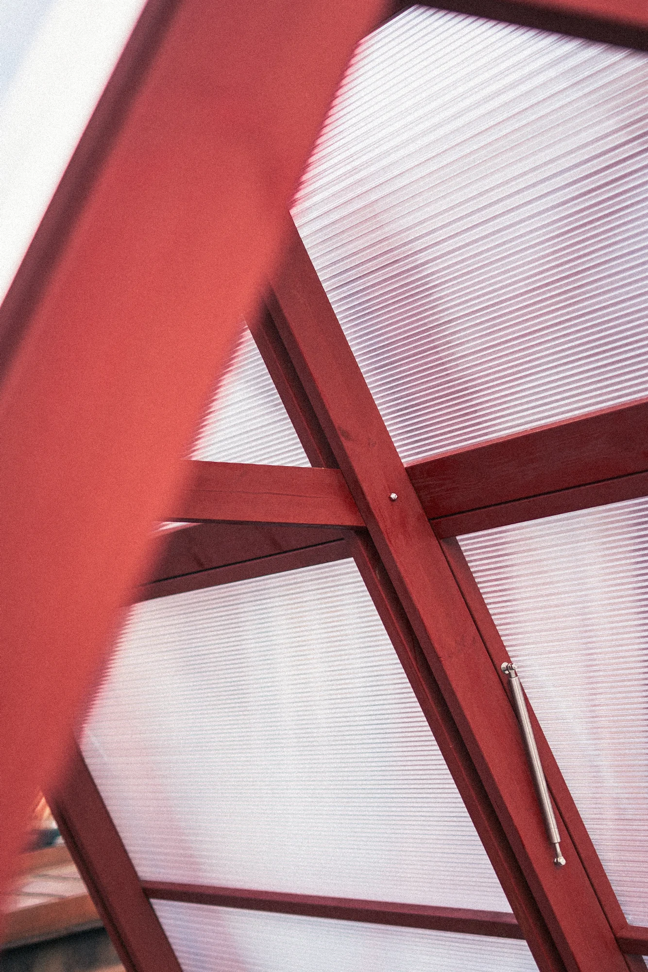 Looking up at the joint where red painted timber beams cross in front of translucent corrugated polycarbonate panels, with a steel gas strut fixed alongside