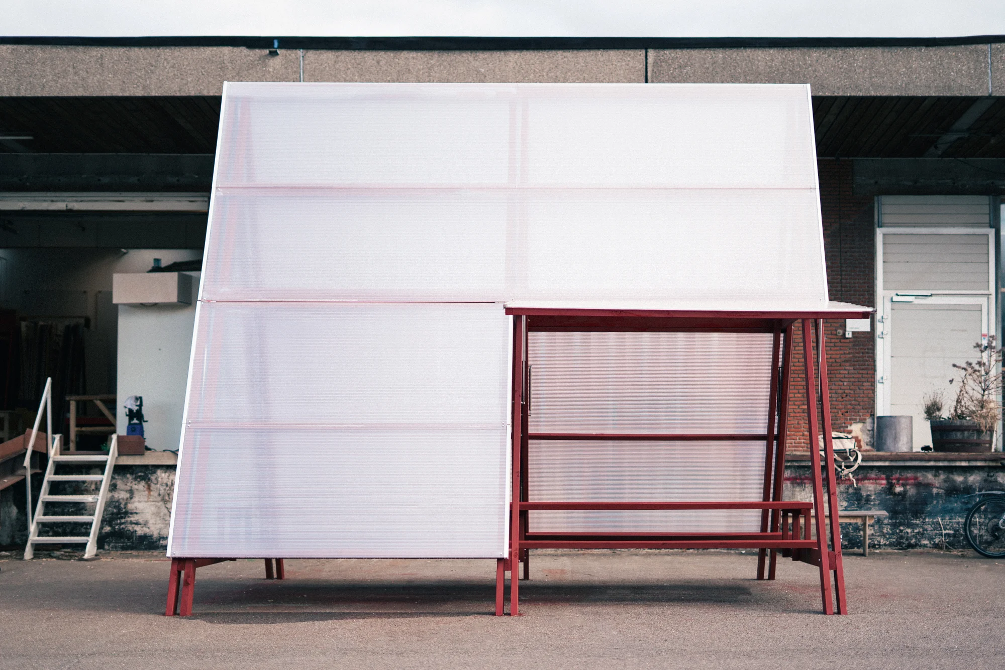 The pavilion seen from the side outside a workshop building: a red timber frame carrying a large sloping roof of translucent corrugated panels, with a bench beneath the open end