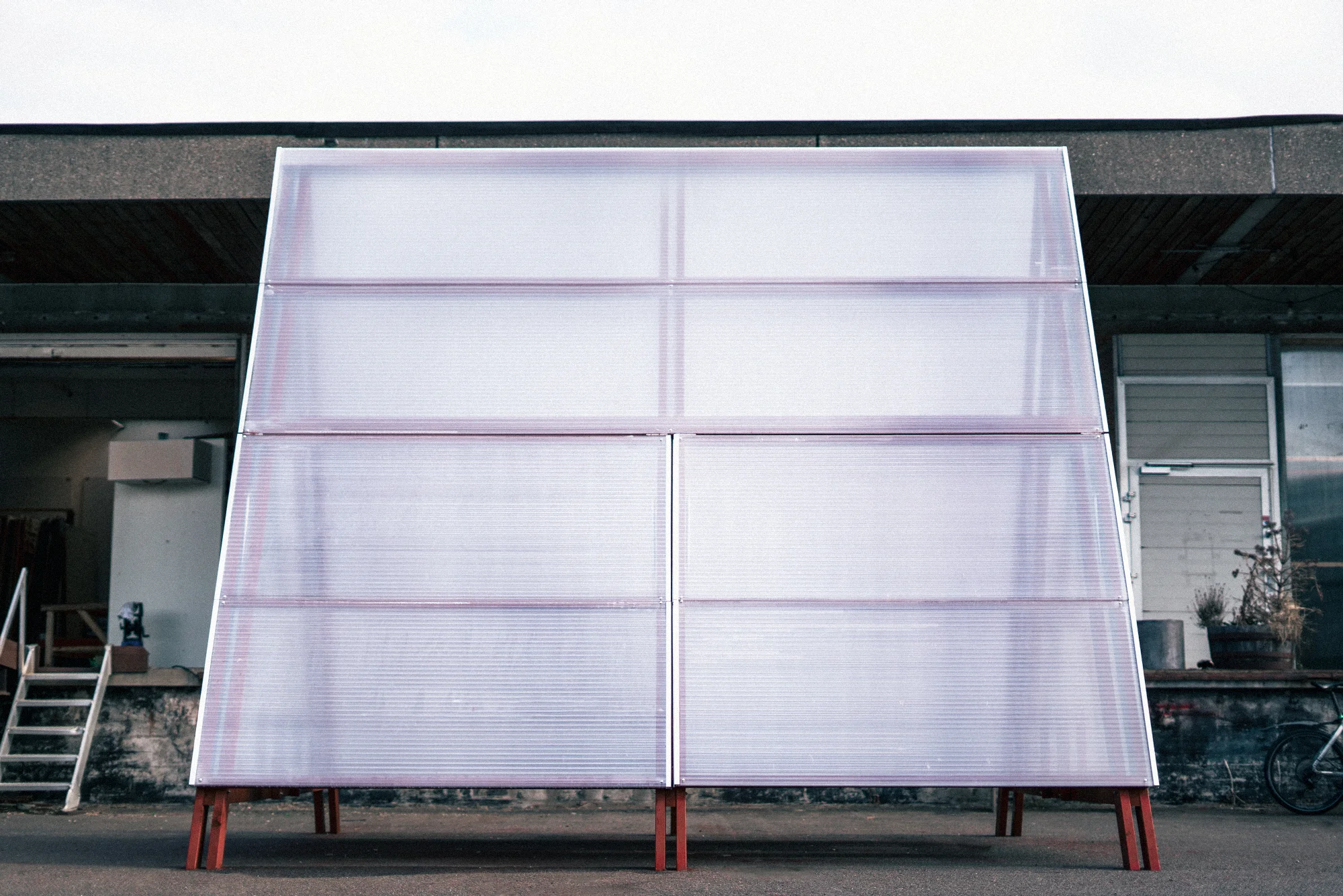 The pavilion head on with every panel closed, the translucent corrugated sheets forming one large sloping face above short red legs