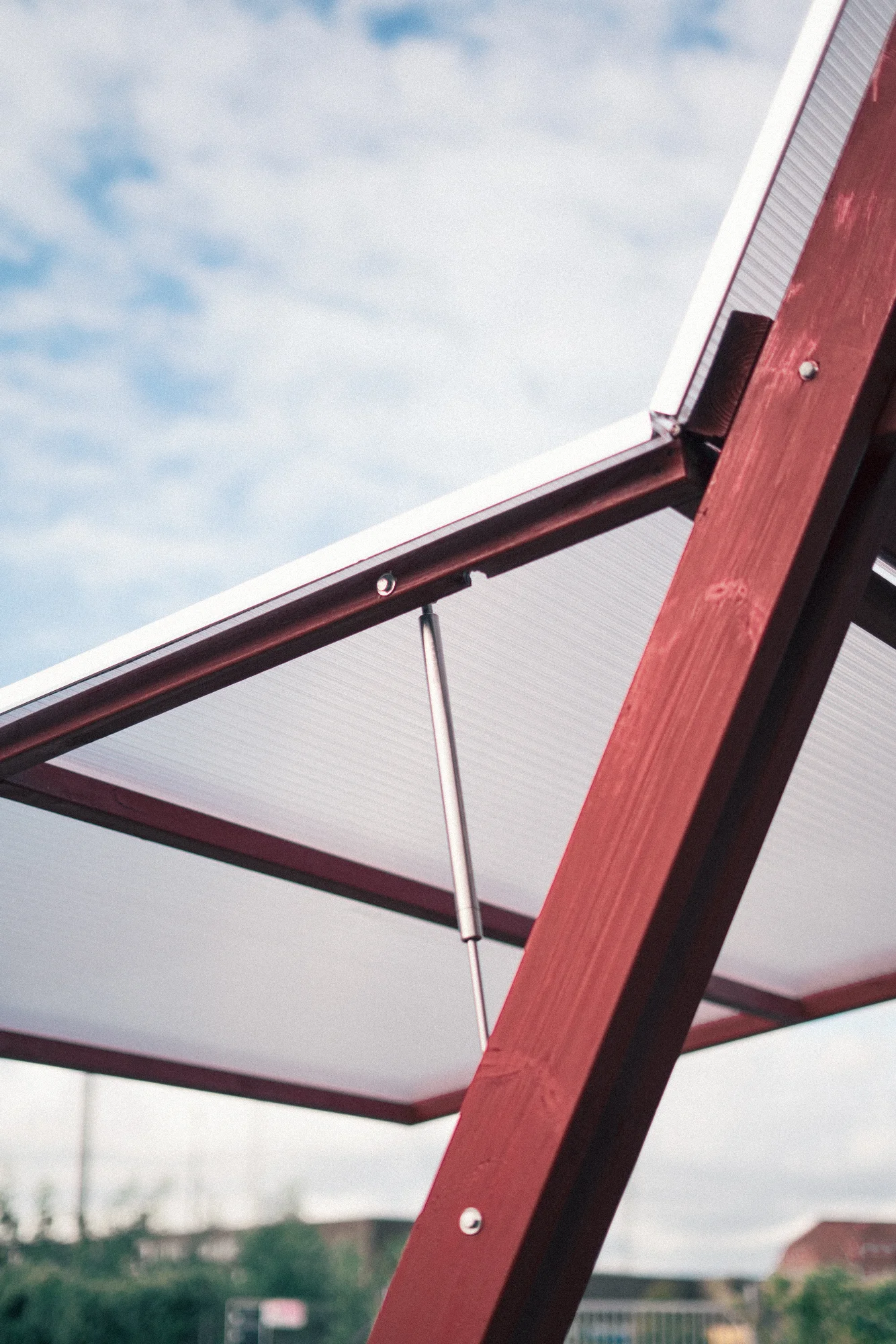 Looking up from underneath at a raised translucent panel held open by a steel gas strut on the red timber frame, against a cloudy sky
