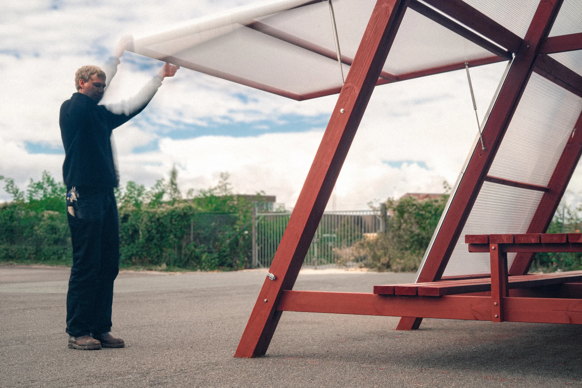 A person lifting one of the translucent panels open by hand on the red timber pavilion, with the long red bench and table visible beneath