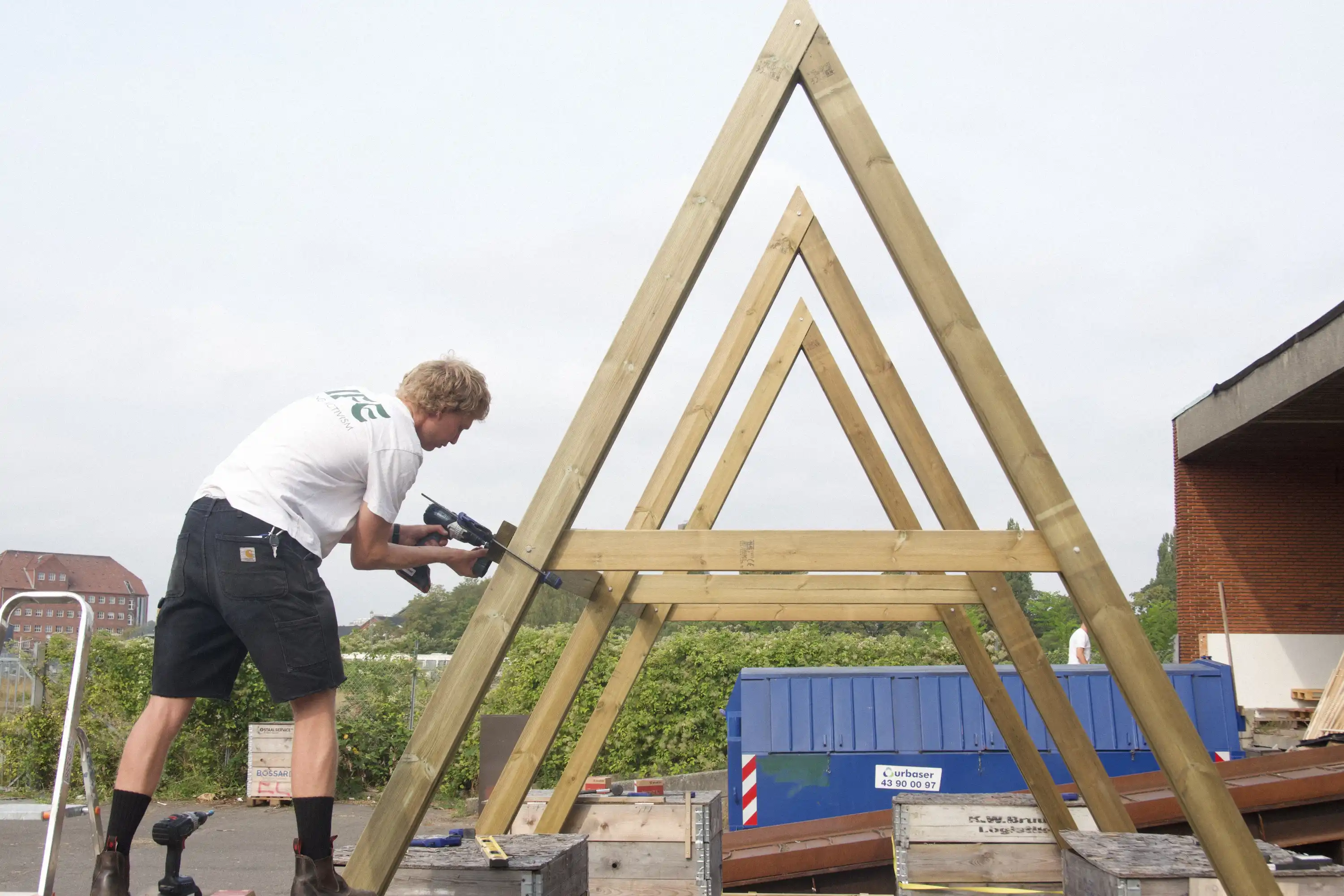 A person drilling a cross beam into three nested triangular timber A-frames standing in a yard, the structure still unpainted