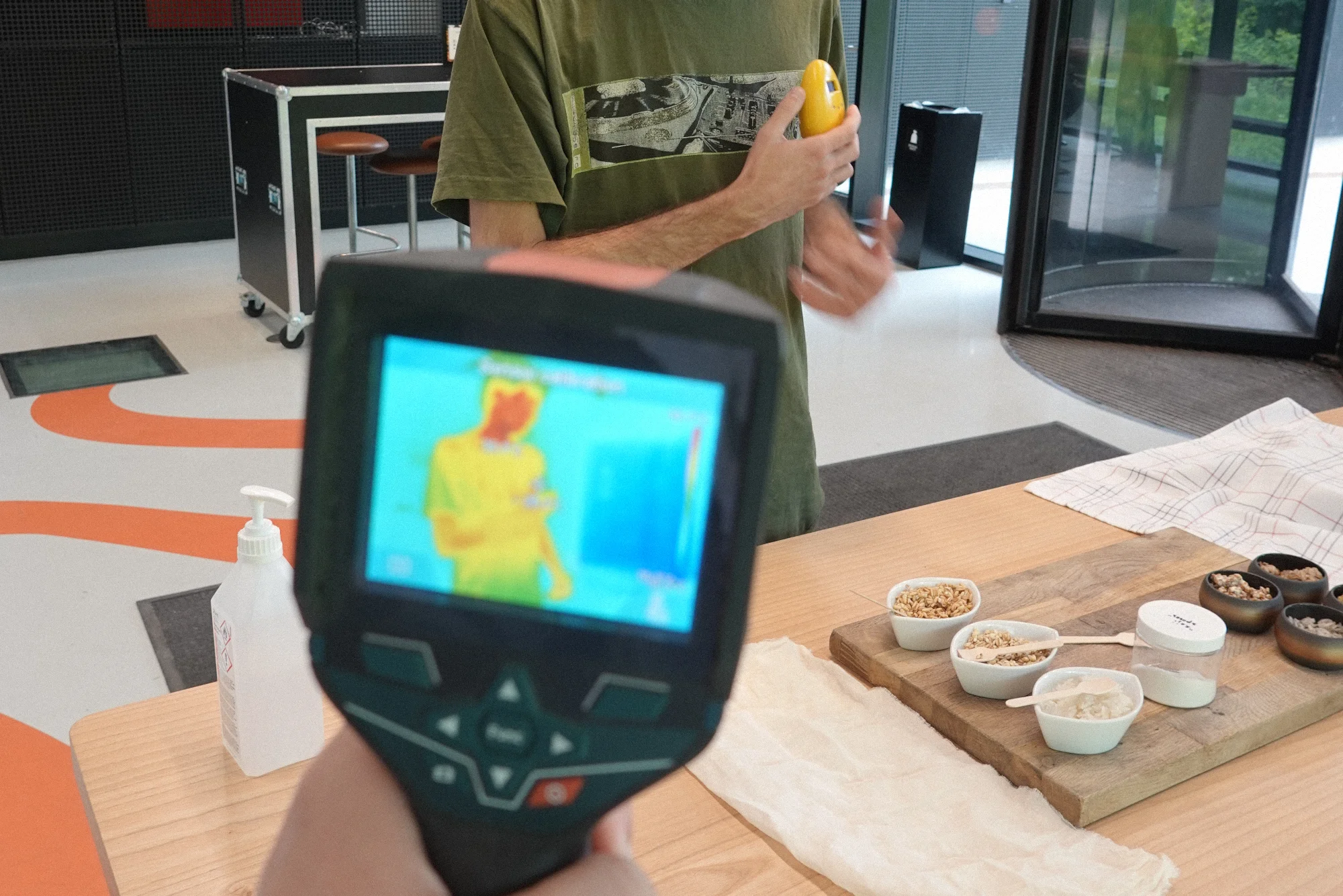 A thermal camera screen showing the body heat of a person holding the device against their chest, with bowls of rice and koji grains on a wooden board beside them