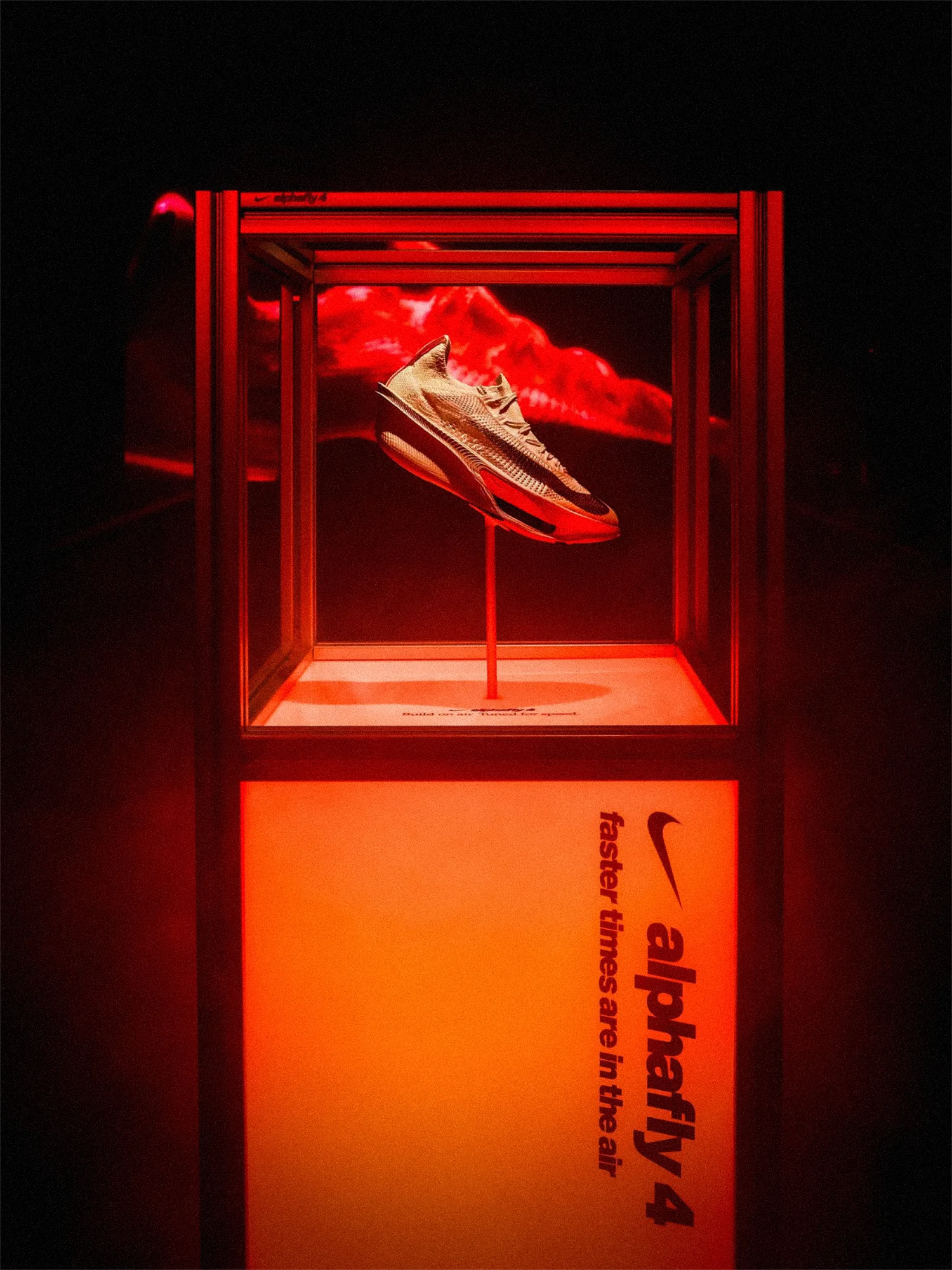 Nike Alphafly 4 running shoe suspended on a thin rod inside a lit glass case, a red cloud form behind it, on a plinth glowing from within with the Nike swoosh and the words alphafly 4, faster times are in the air